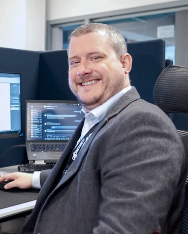 A smiling man seated in front of a computer with multiple screens displaying code, dressed in business attire with a grey blazer and a blue tie, in an office setting