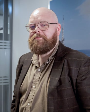 A bearded man with glasses, wearing a brown jacket and a khaki shirt, giving a serious look, with an office background