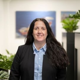 A professional woman with long hair, wearing a black blazer and a blue shirt, smiling at the camera in an office environment with pictures of ships and green plants in the soft-focused background
