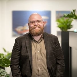 A man with a beard and glasses, smiling at the camera, wearing a brown jacket over a beige shirt, with an office environment including green plants and maritime-themed pictures in the background.