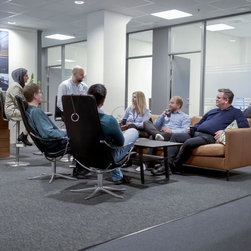 A group of professionals seated in a circle in an office setting, engaged in a discussion. Some members are on couches while others are on office chairs, creating an informal and collaborative atmosphere.