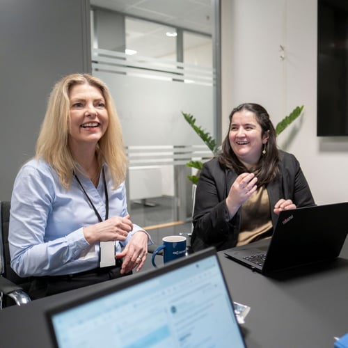 Two smiling women in a professional office environment, one holding a mug, engaged in a cheerful conversation with a laptop open in front of them, indicating a collaborative work atmosphere.
