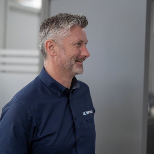 A profile view of a smiling man with gray hair, wearing a dark blue shirt. The shirt has a logo on the chest, and the man appears to be in a well-lit, indoor environment with a door in the background.