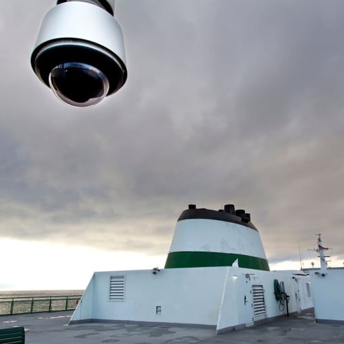 The deck of a ship featuring a large security camera in the foreground, with the ship's funnel and part of the bridge visible in the background under an overcast sky.