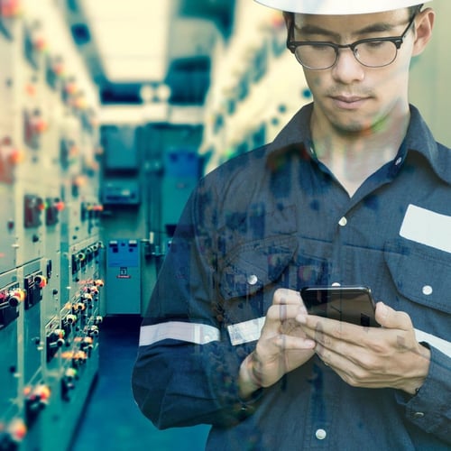 A man in a work shirt and safety helmet focused on his smartphone, with an industrial setting blurred in the background featuring rows of electrical panels and controls
