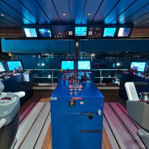 Control room of a modern ship with an array of electronic screens and navigation equipment. The room is lit with ambient blue lighting and has a central console with various control buttons and joysticks, with chairs and workstations arranged around it