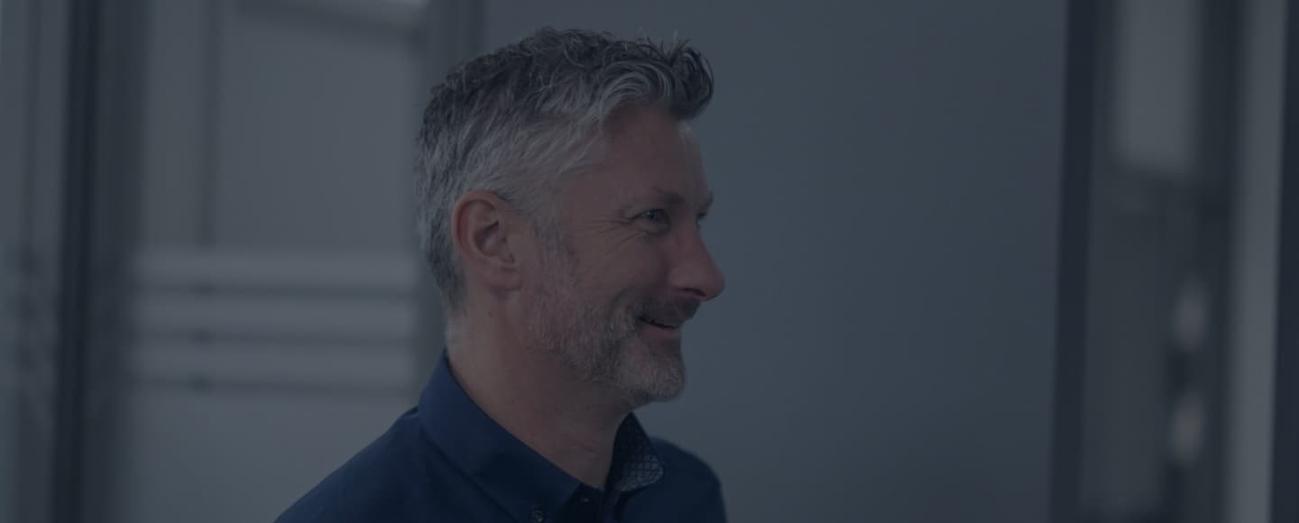 Profile view of a smiling man with gray hair, in a well-lit room. He's wearing a dark blue shirt with a visible collar of a different pattern underneath. The background is out of focus, with neutral colors suggesting an interior setting.