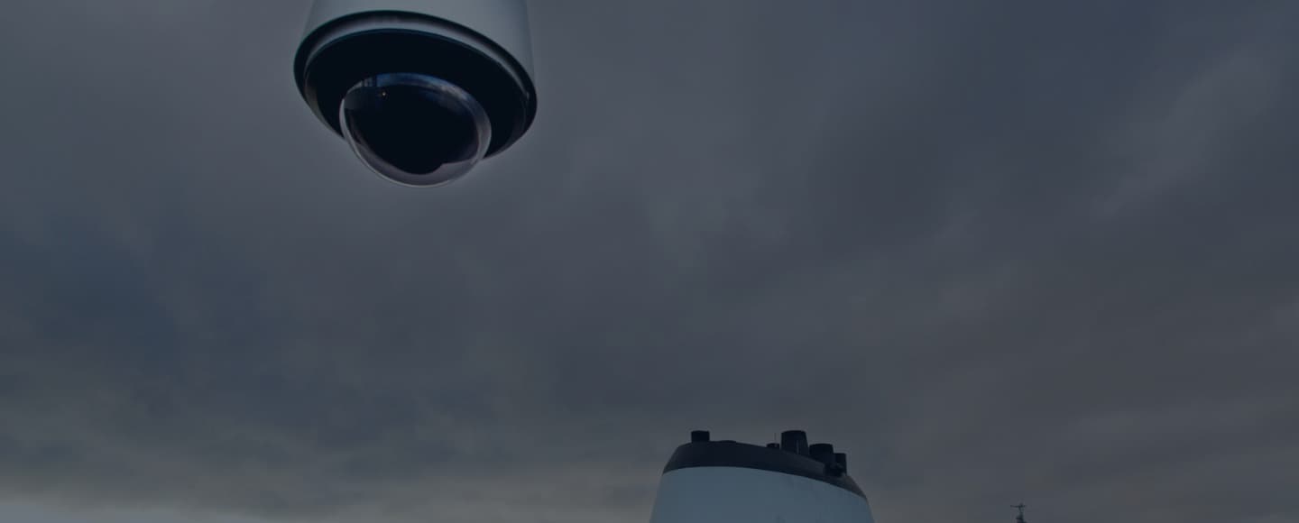 A low angle view of a surveillance camera hanging overhead against a cloudy sky, with the silhouette of a ship's funnel also visible in the frame. The image conveys a sense of monitoring and security on a ship, with a focus on the camera's presence against the overcast weather conditions