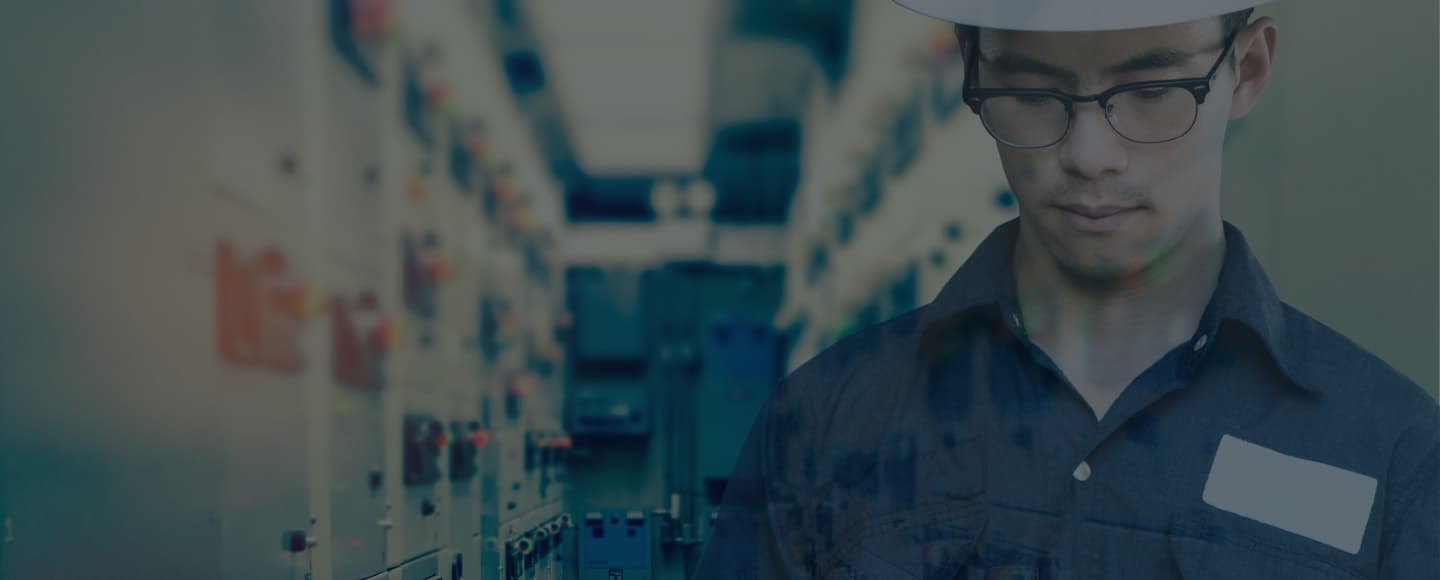 A banner image featuring a man in the foreground wearing safety glasses and a dark blue shirt with a name tag, looking down thoughtfully. The background is blurred but suggests an industrial setting with a long corridor flanked by machinery or equipment, reflecting a cool blue tone overall