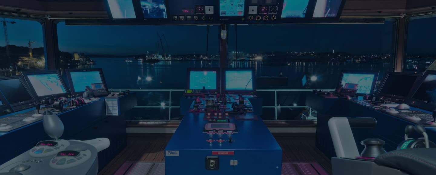 Night view inside a ship's bridge, with control panels and electronic navigational equipment. The bridge is lit by the glow of multiple monitors and the ambient light from outside, with a panoramic window showing a harbor with lights reflecting on the water.