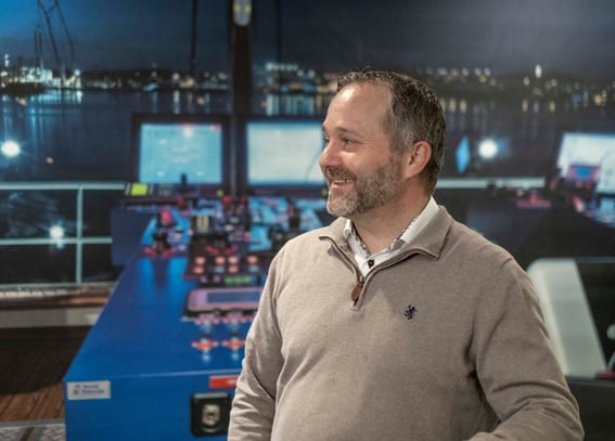 A smiling man in a casual grey pullover with a polo logo stands in a room with a view of a harbor at night through large windows. Behind him is an illuminated control room with multiple computer screens and control panels
